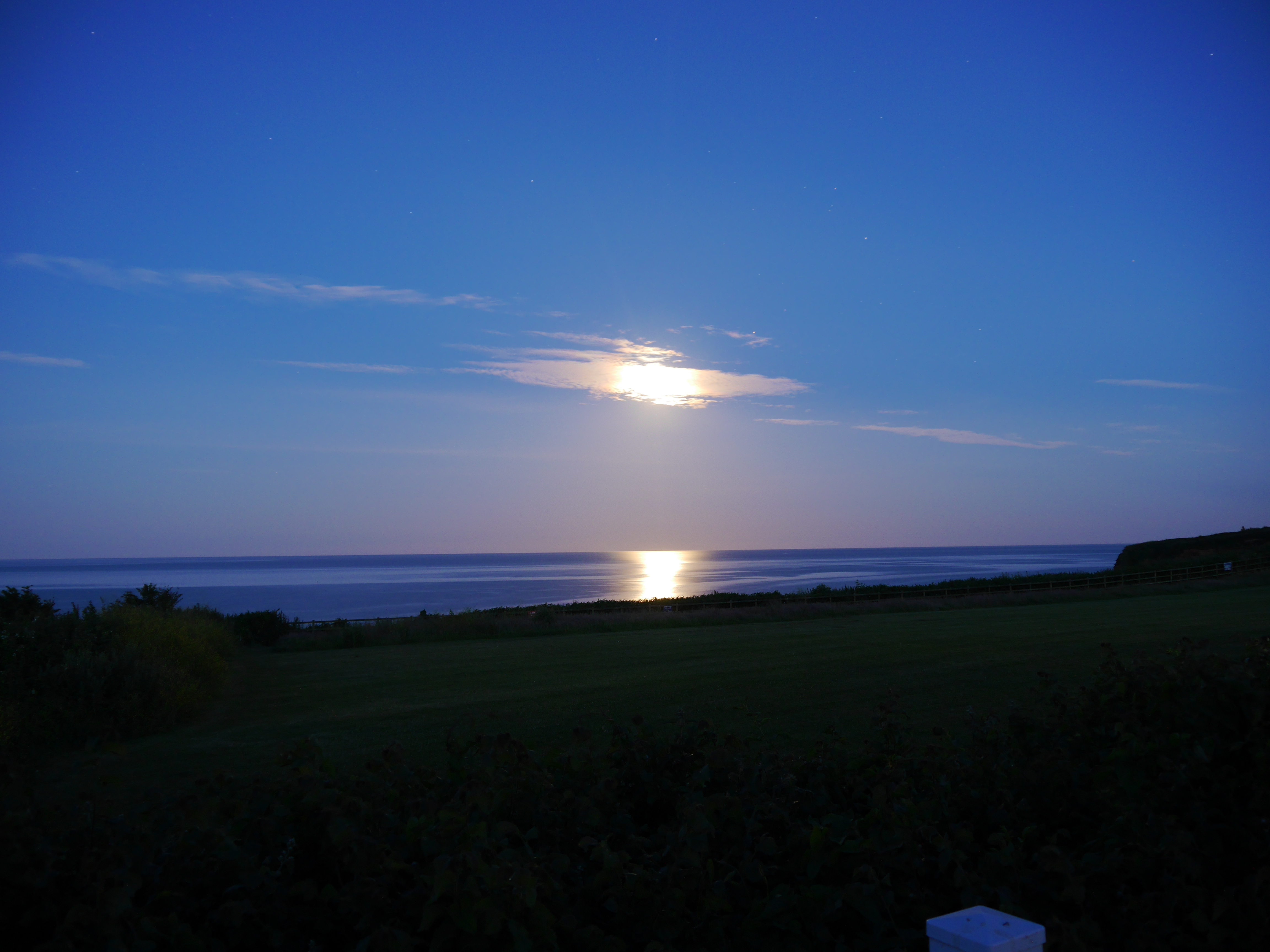 A photograph of the night sky. The moon is partially obscured by a cloud, and the moonlight reflects off the sea