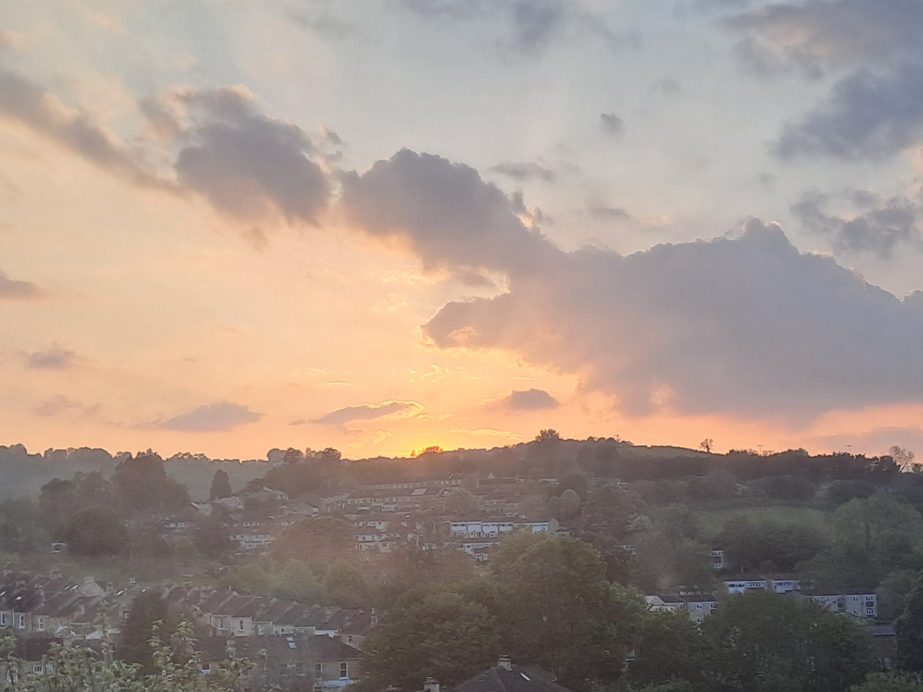 A photograph of the sun setting behind a hill, with bright orange rays of light shining between the clouds on the pale blue sky