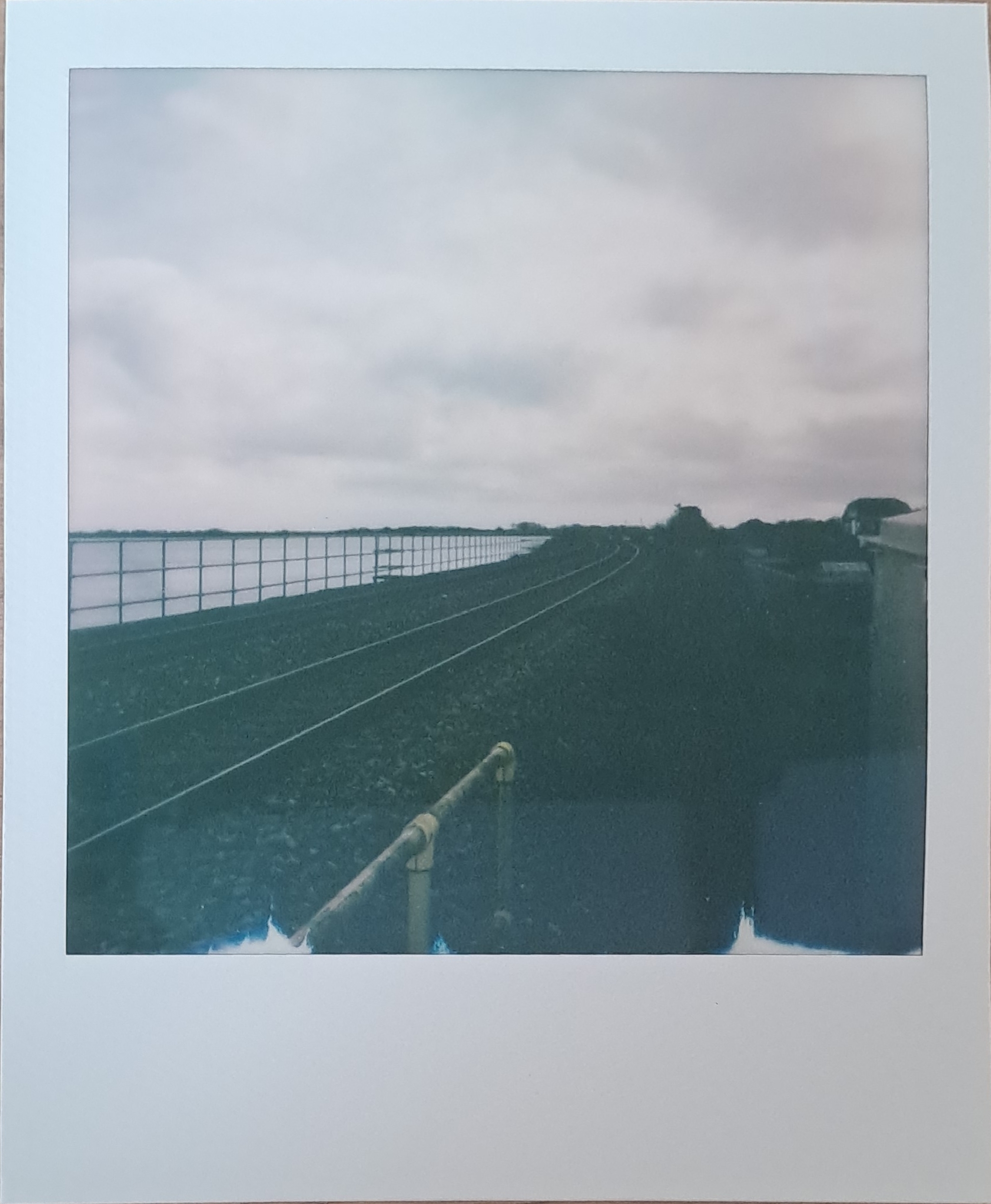 A Polaroid of a railway, with a metal railing, along the bank of a lake